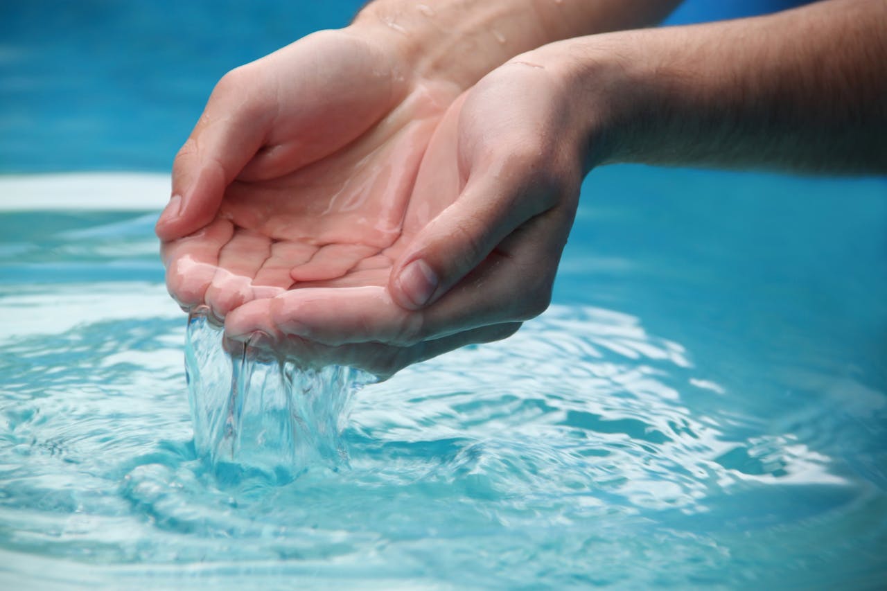 Close-up of hands scooping clear blue water from a reflective pool outdoors.